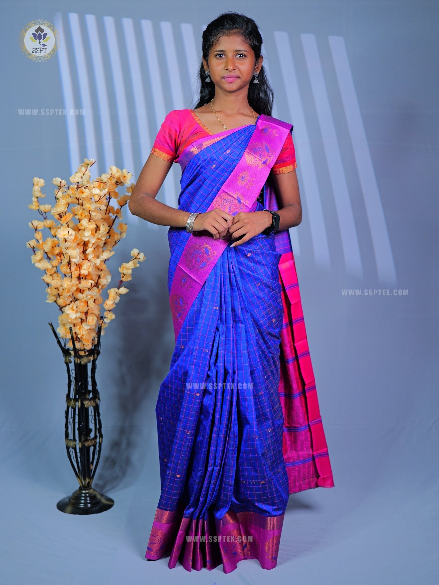 A woman wearing a black and purple sari with a green border, posed in front of a light blue studio backdrop with a decorative vase of yellow flowers.